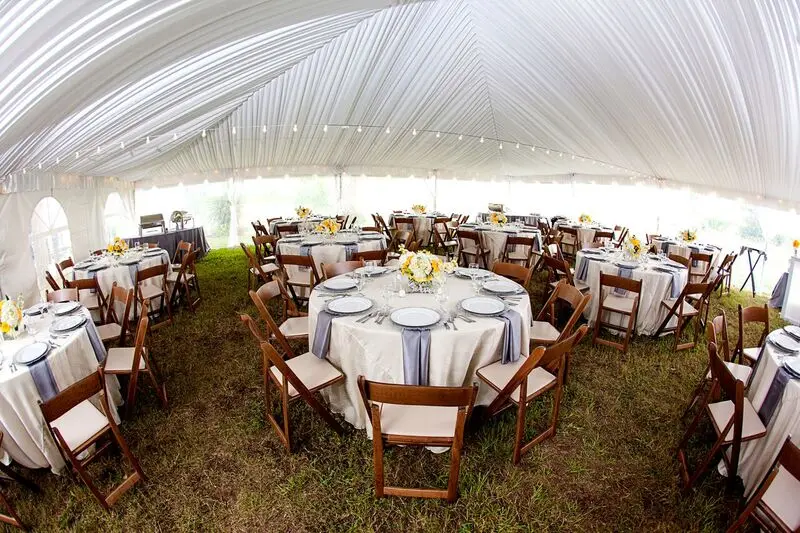 A tented wedding reception set by All About Events, with round tables, wooden folding chairs, string lights and a draped tent ceiling over grass