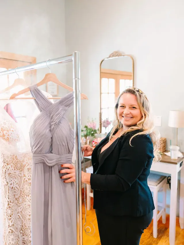 Brittany Alphonso of Honey Bee Events, smiling beside a rack of wedding and bridesmaid dresses