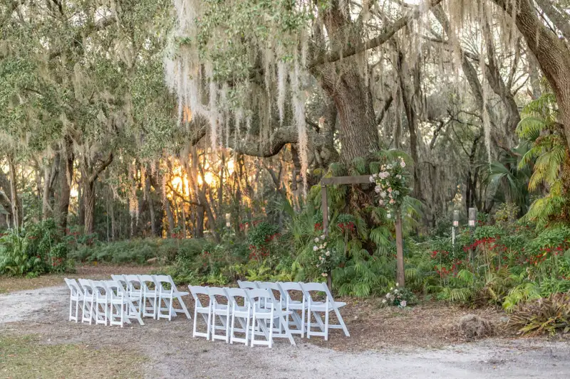 White folding chairs facing a wooden arbor with blush florals under moss-draped oaks at sunset at Fern Oak Estate in Pomona Park Florida