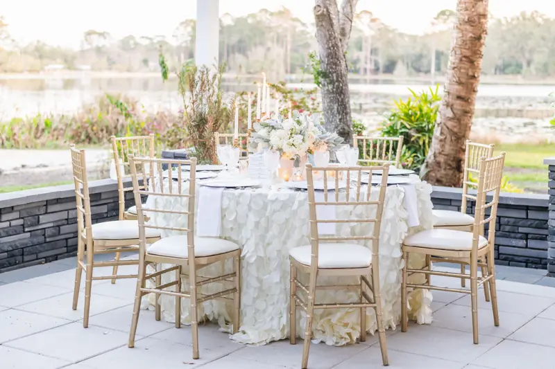 Reception table set with candles and gold chairs on the covered terrace at Fern Oak Estate at dusk, palms and the lake behind