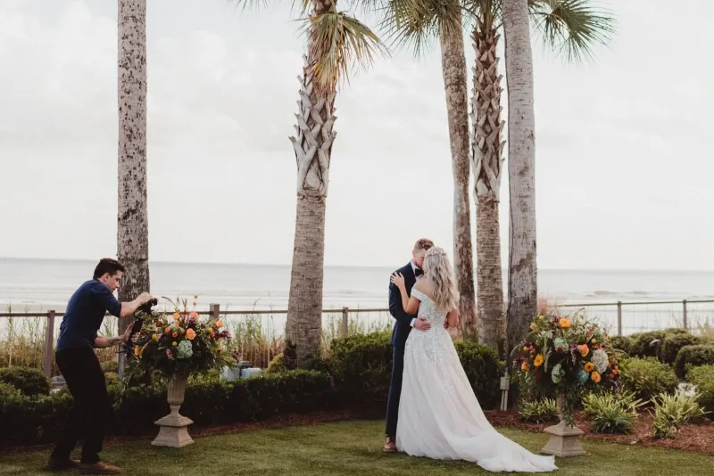 A Rene Visuals cinematographer filming a couple at an oceanfront Florida wedding ceremony framed by palm trees