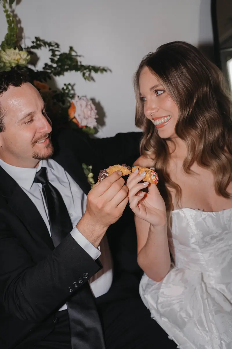A bride and groom holding up two filled cannolis, one topped with chocolate chips and one with rainbow sprinkles
